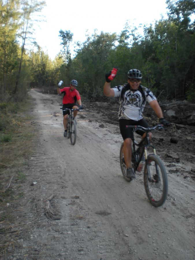 Two mountain bikers riding on a dirt trail surrounded by trees. One rider is wearing a red shirt and raising a can, while the other is in a black and white jersey, smiling and holding a drink. Oleta River State Park mountain bike trail.