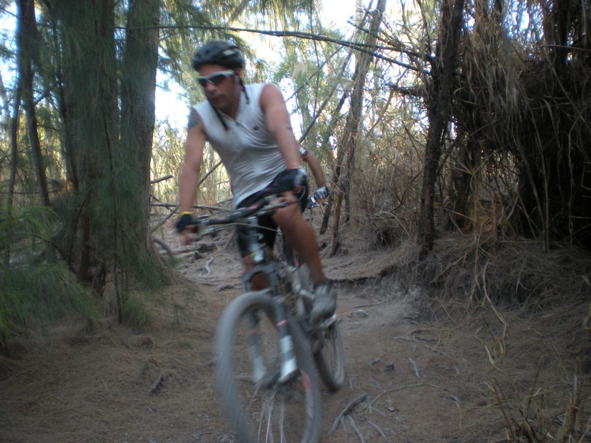 A mountain biker riding along a narrow dirt trail surrounded by trees, with a blurred background suggesting speed. The cyclist is wearing a helmet, sunglasses, and a sleeveless shirt, navigating through a natural setting. Oleta River State Park mountain bike trail.