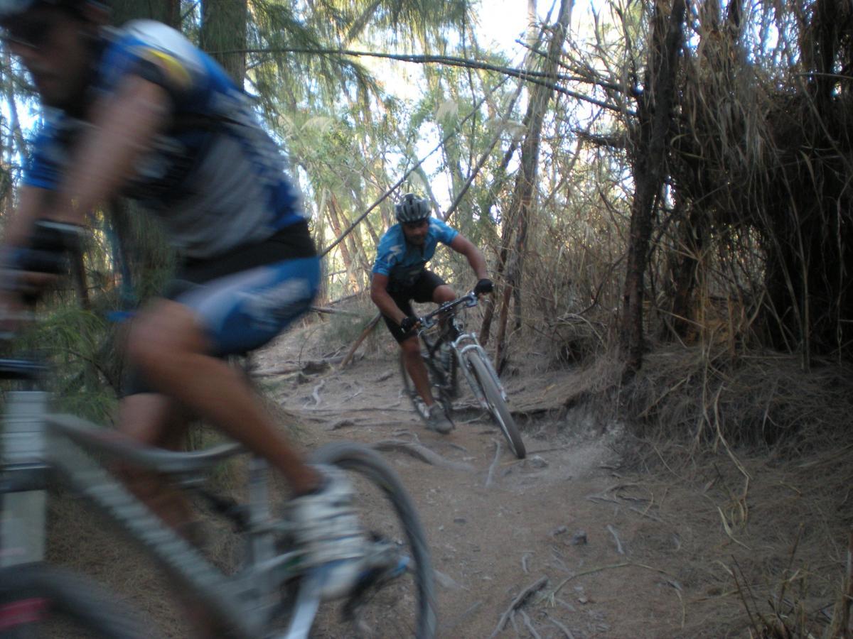 Two mountain bikers riding through a wooded trail, one in motion and slightly blurred, while the other is focused and closer to the camera. The path is surrounded by trees and underbrush, suggesting a rugged outdoor environment. Oleta River State Park mountain bike trail.