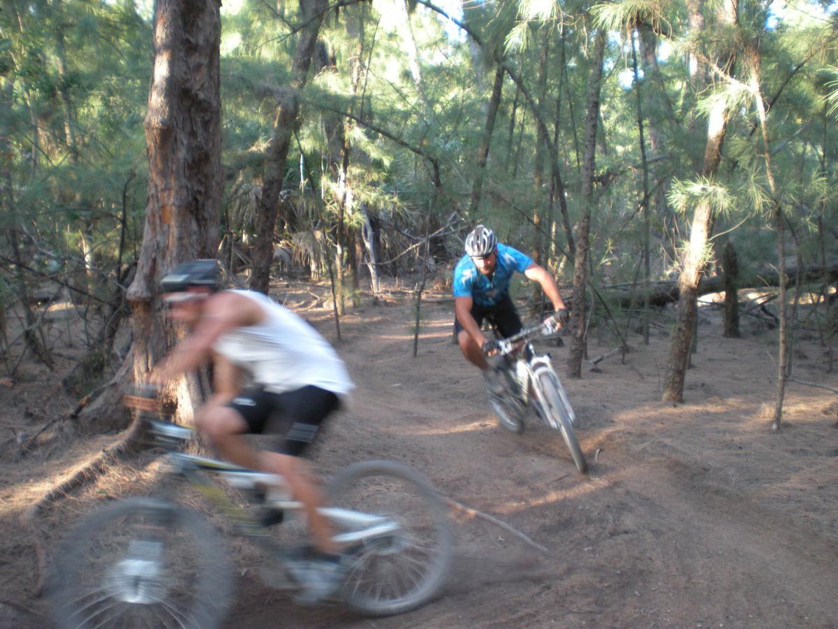 Two mountain bikers navigating a sandy trail in a wooded area. One rider in a blue shirt is in focus and leaning into a turn, while the other, dressed in a white tank top, is slightly blurred as they pass by. The surrounding forest includes tall trees and underbrush, suggesting an adventurous outdoor setting. Oleta River State Park mountain bike trail.