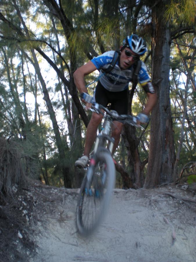 A mountain biker in mid-air as he jumps over a sandy path, surrounded by trees in a natural setting. The cyclist is wearing a blue jersey, cycling shorts, and a helmet, with a determined expression on his face. Oleta River State Park mountain bike trail.