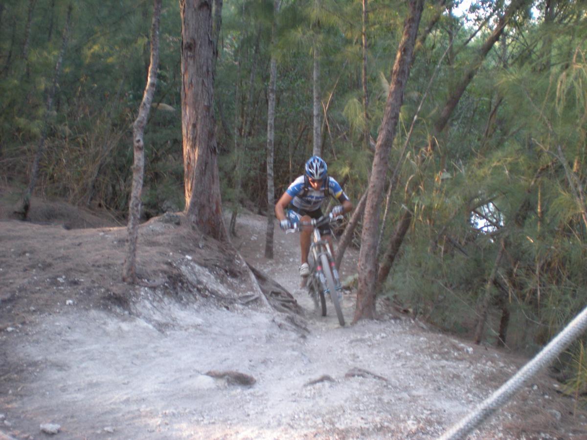 A mountain biker navigates a rocky trail surrounded by trees, wearing protective gear and focused on the path ahead. The dirt trail is winding and dappled with sunlight filtering through the foliage. Oleta River State Park mountain bike trail.