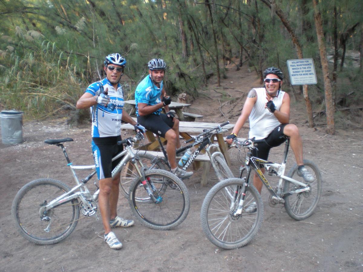 Three mountain bikers pose for a photo in a wooded area. Each wears cycling gear and helmets, with two men giving a thumbs-up and one smiling broadly. They are leaning against their bicycles, which rest on the dirt ground near a picnic table. In the background, there is a sign that encourages volunteering for trail maintenance. Lush greenery surrounds the scene. Oleta River State Park mountain bike trail.