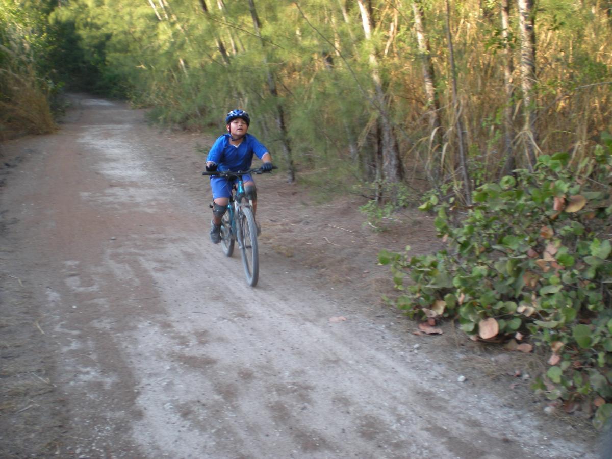 A young child riding a bicycle on a dirt path surrounded by greenery and trees. The child is wearing a blue outfit and a helmet. The scene captures a moment of outdoor activity in a natural setting. Oleta River State Park mountain bike trail.