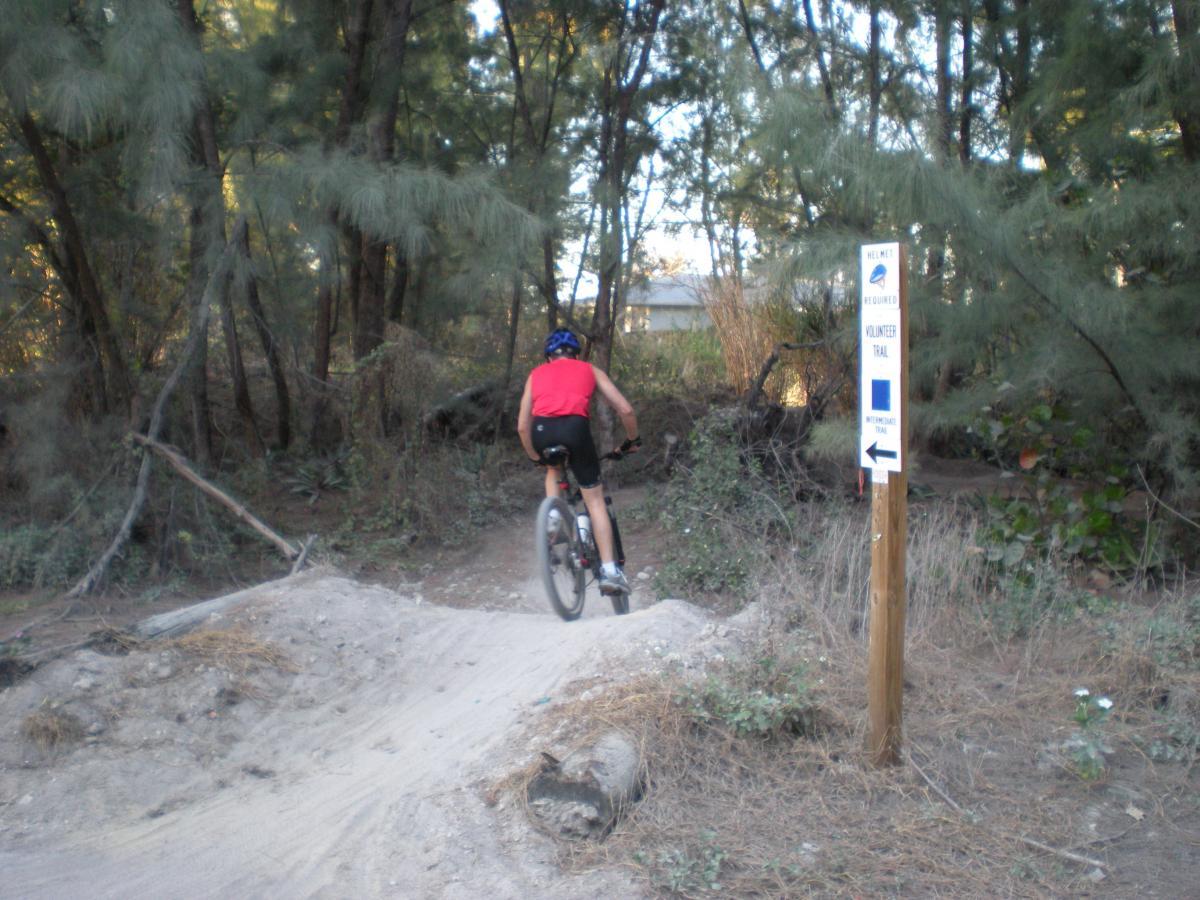 A person in a red shirt and helmet riding a mountain bike on a dirt trail, surrounded by trees. A trail sign indicating a "Volunteer Trail" with a blue marking is visible on the right. Oleta River State Park mountain bike trail.