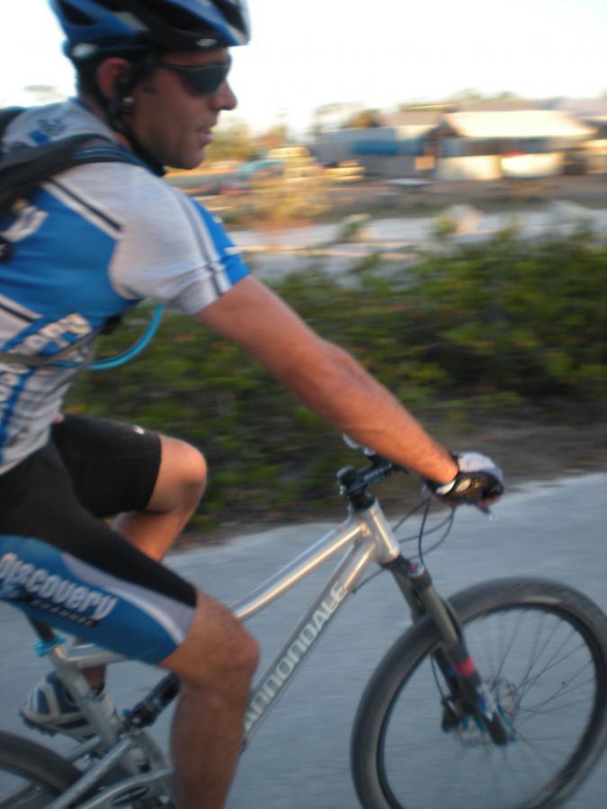 A cyclist wearing sunglasses and a blue and white cycling jersey rides a silver mountain bike on a path, with greenery and buildings in the background. The image captures the motion of the cyclist in an outdoor setting during daylight. Oleta River State Park mountain bike trail.