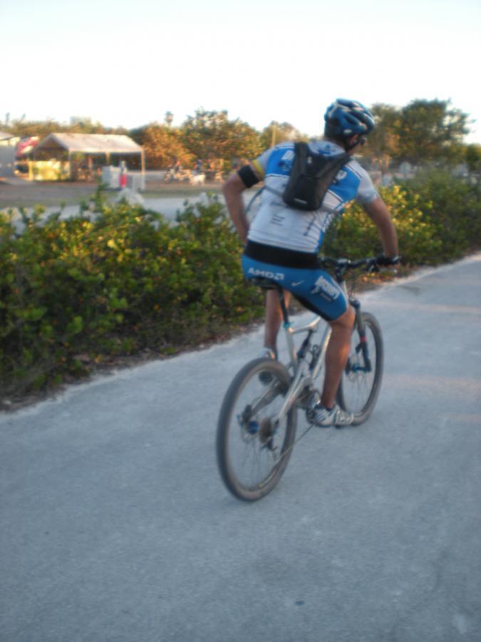 A cyclist wearing a blue and white jersey rides a mountain bike along a paved path surrounded by greenery. The scene depicts a leisurely outdoor biking activity during sunset. Oleta River State Park mountain bike trail.