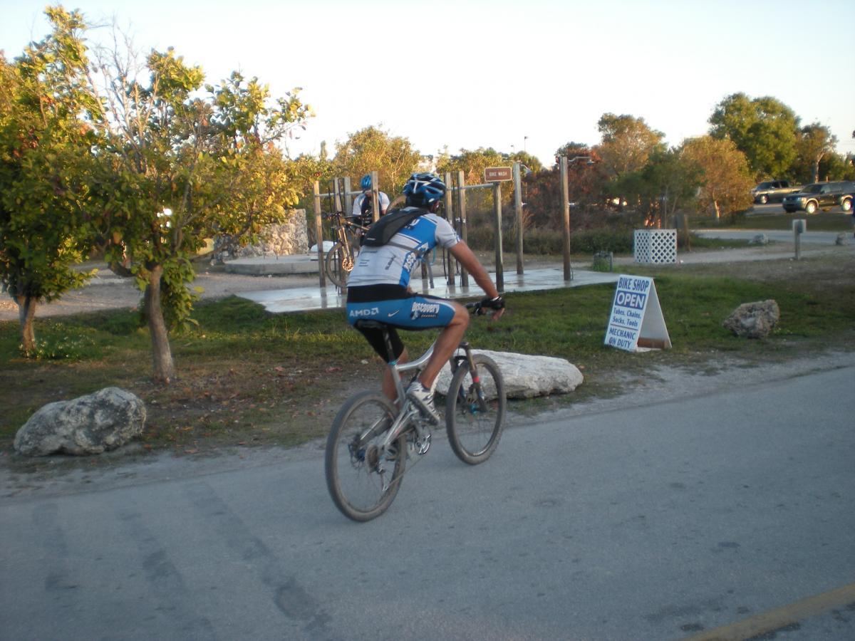 A cyclist in a blue and white jersey rides a mountain bike along a road. In the background, there are trees, a bike repair area, and a sign indicating that a bike shop is open with a mechanic on duty. The scene is set in a park-like environment during the evening. Oleta River State Park mountain bike trail.