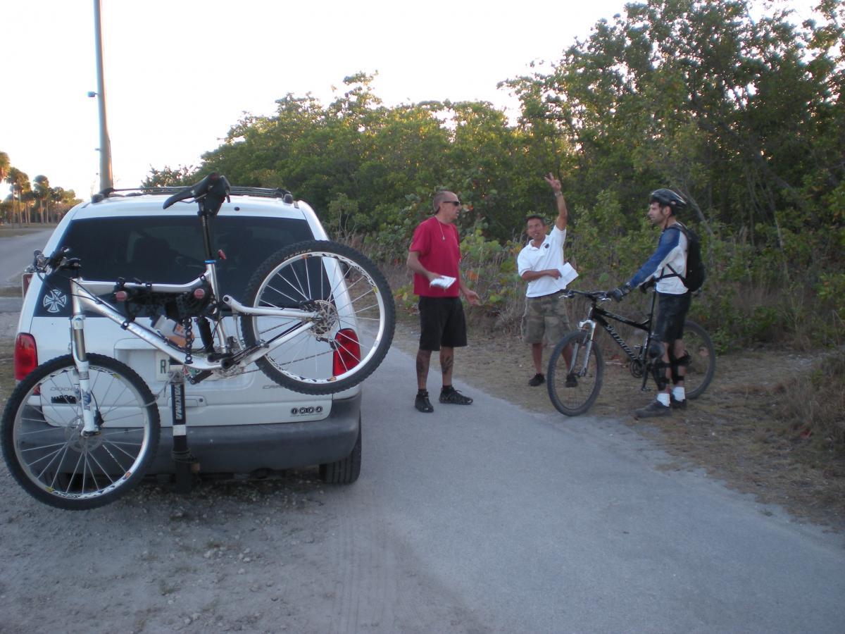 Three individuals are gathered by the side of a dirt road with a white van parked nearby. One person is leaning against the van, while the other two are standing on the side of the road, one gesturing with their hand. A mountain bike is attached to the back of the van. The surrounding area features shrubs and trees, hinting at an outdoor setting, possibly near a biking or nature trail. Oleta River State Park mountain bike trail.