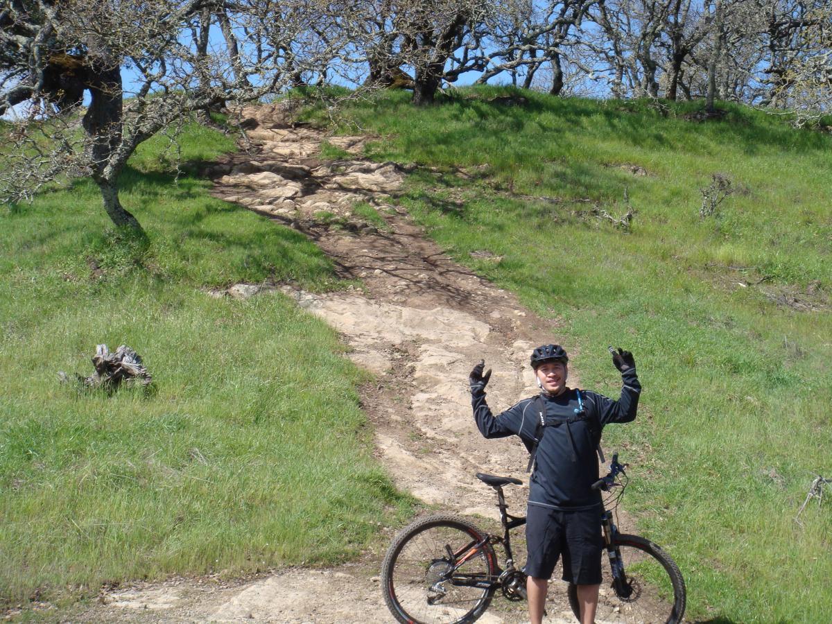 A person in biking gear stands next to a mountain bike on a grassy trail, with rocky terrain leading up a hill. The individual has their arms raised in excitement, and there are trees in the background against a clear blue sky. Rockville Park mountain bike trail.