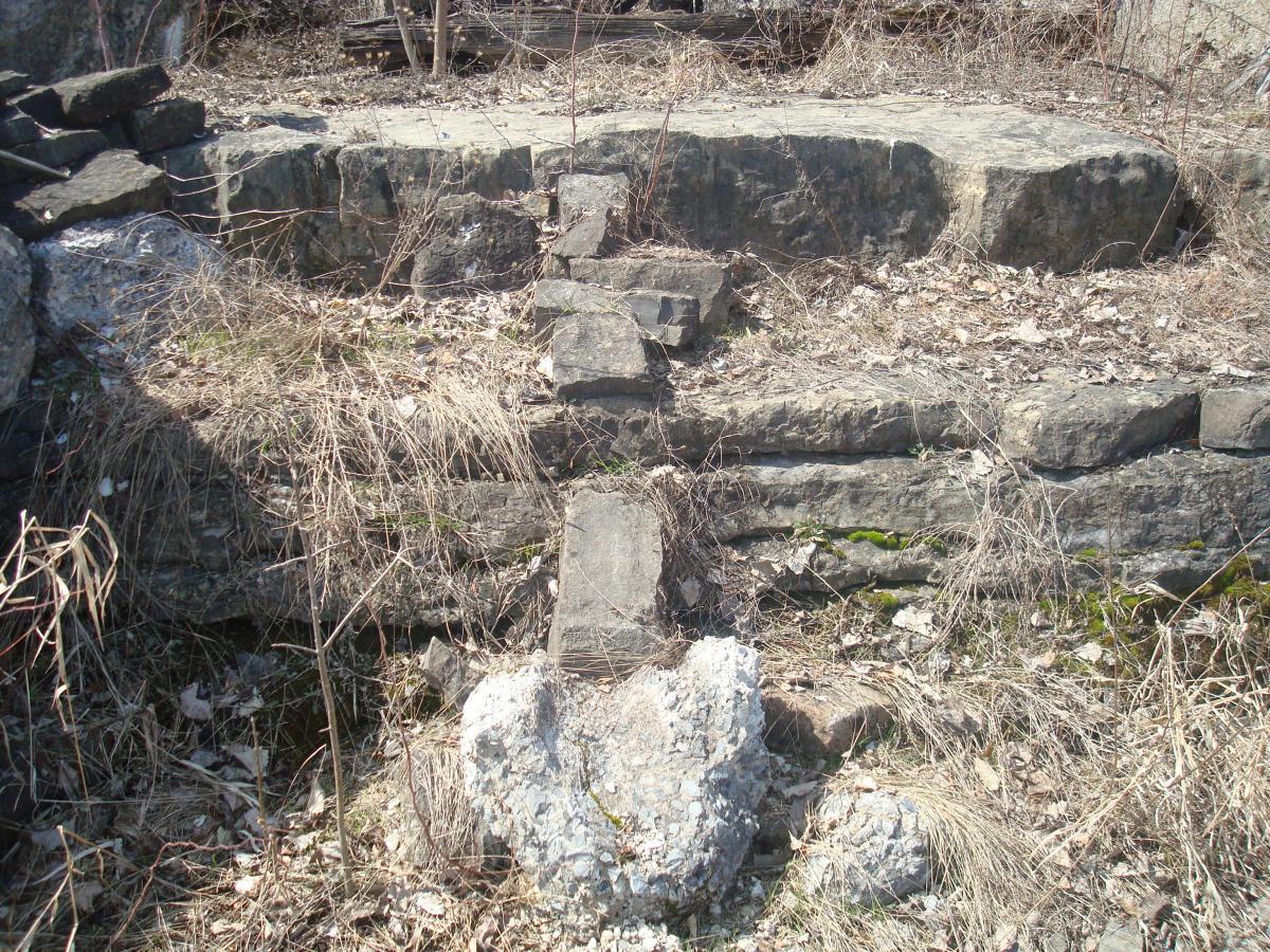 A weathered stone structure overgrown with grass and weeds, featuring large, uneven rock formations arranged in a cross-like shape, surrounded by dry leaves and sparse vegetation. Blue Trail mountain bike trail.
