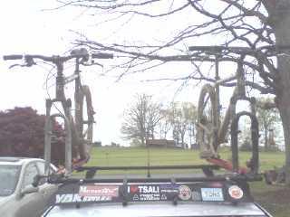 Two bicycles are mounted on a roof rack atop a vehicle, with a view of a grassy area and trees in the background. The image has a cloudy sky and features various stickers on the roof rack. Tsali Right Loop mountain bike trail.