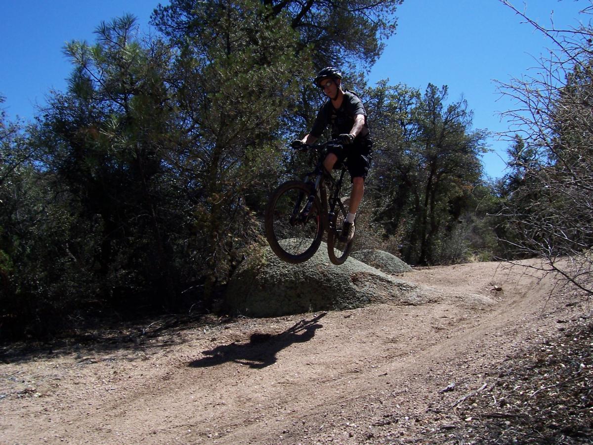 A mountain biker mid-air, performing a jump over a rocky outcrop on a dirt trail, surrounded by trees under a clear blue sky. Granite Basin Loops mountain bike trail.
