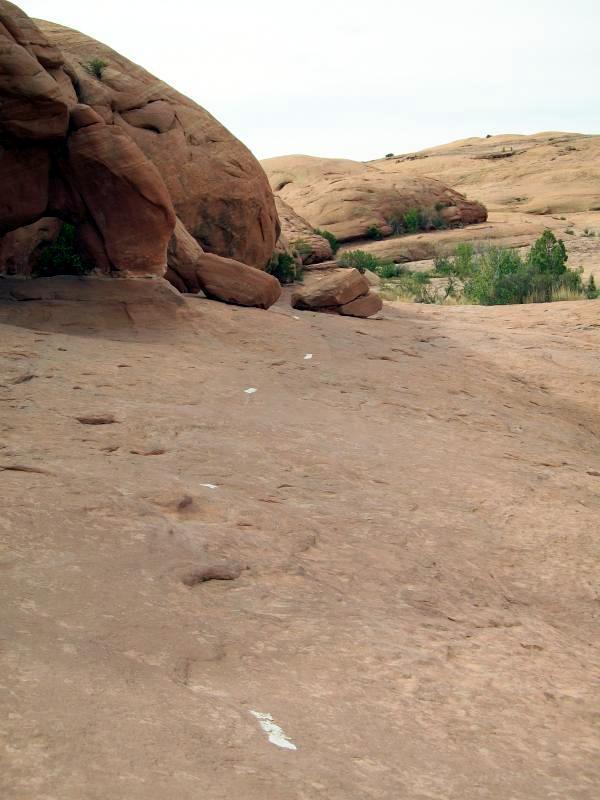 A rocky landscape with large sandstone formations and a smooth, dusty path. Sparse vegetation can be seen in the background, and white markings are visible on the ground, suggesting a trail or pathway. The sky is overcast with a light gray hue. Slickrock mountain bike trail.