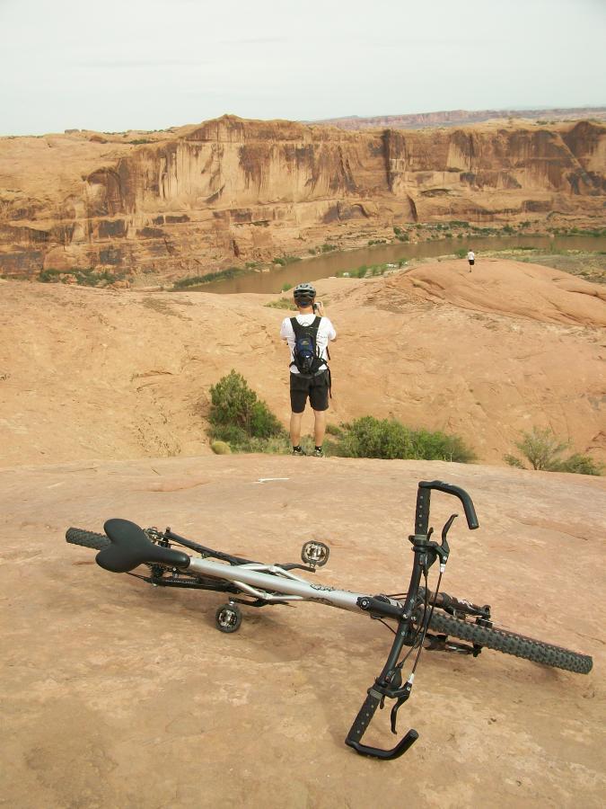 A mountain biker's bicycle lies on a rocky ledge overlooking a canyon, while a person stands in the background, taking a photo of the scenic view. Lush green vegetation can be seen along the river winding through the canyon below, framed by the reddish sandstone formations. Slickrock mountain bike trail.