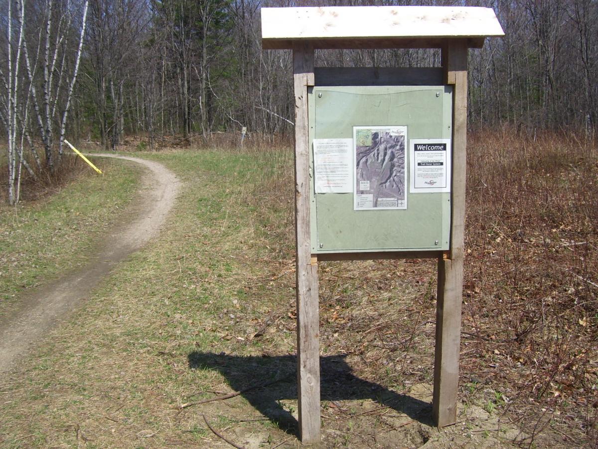 A wooden information board displaying maps and guidelines stands at the entrance of a dirt path winding through a natural landscape. The path leads into a wooded area with sparse vegetation, surrounded by bare trees and green grass. Sunny Hollow mountain bike trail.