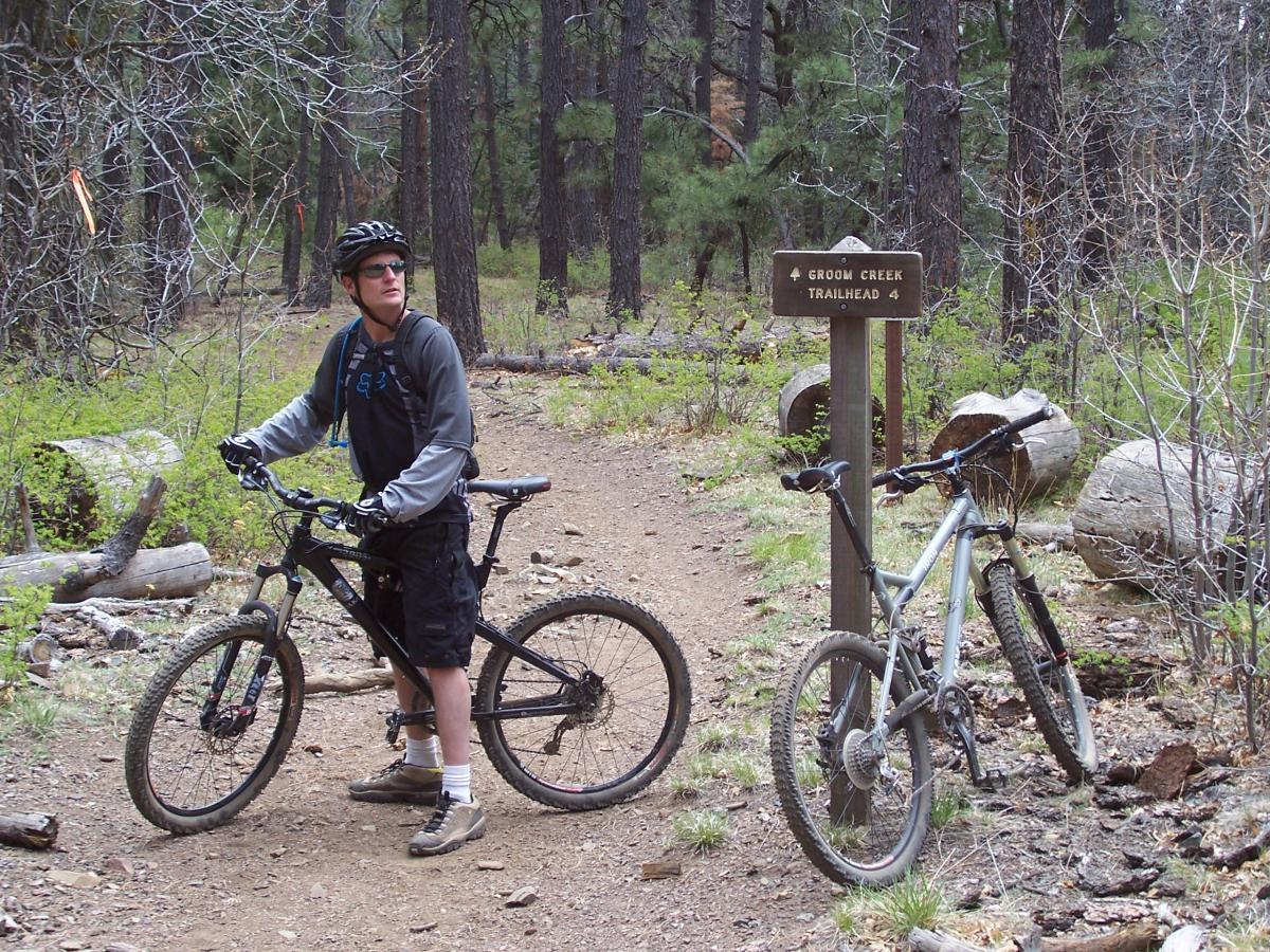 A mountain biker wearing a helmet and sunglasses stands on a dirt trail, looking towards a sign marked "Groom Creek Trailhead." A second bike leans against the sign. The surrounding area is forested with tall trees and scattered logs. Groom Creek Loop mountain bike trail.