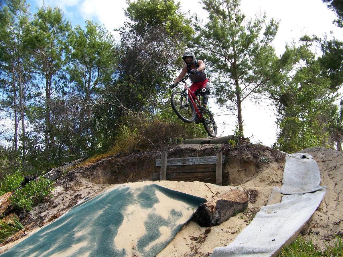 A mountain biker performing a jump over a sandy ramp, surrounded by trees and foliage. The biker is wearing a helmet and protective gear, showcasing an action-packed moment in a natural outdoor setting. Jonathan Dickinson State Park mountain bike trail.