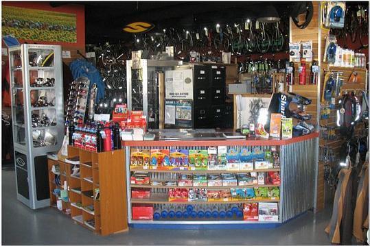 Image of a retail store interior featuring a checkout counter with various products displayed. The counter is surrounded by racks of merchandise, including cycling gear and accessories. The background shows a collection of bicycles and sports equipment, creating a vibrant shopping atmosphere.