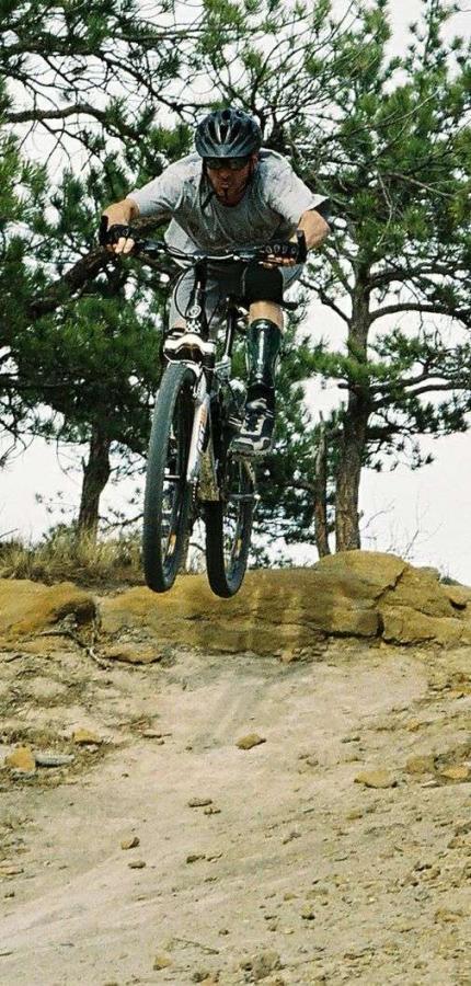 A mountain biker in mid-air performs a jump over a rocky terrain, surrounded by trees and a natural landscape. The cyclist is wearing a helmet and protective gear, showcasing an active and adventurous moment in mountain biking. Palmer Park mountain bike trail.