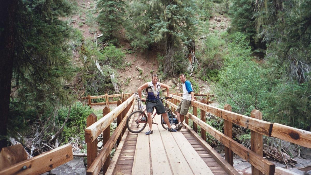 Two cyclists stand on a wooden bridge surrounded by lush greenery. One cyclist is leaning against the railing with a mountain bike beside him, while the other poses confidently with his bike. The scene captures a moment of camaraderie in a natural outdoor setting. Hermosa Creek Trail mountain bike trail.