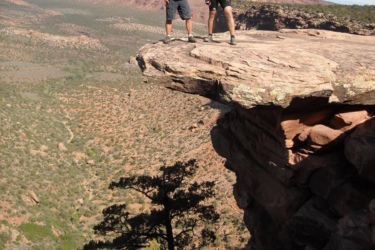 Two individuals standing on a rocky ledge overlooking a vast landscape of hills and valleys. They are wearing casual hiking attire and helmets, with one person in a tank top and the other in a red shirt. The scene features a clear blue sky and a rugged terrain filled with greenery and rock formations in the background. Kokopelli Trail mountain bike trail.