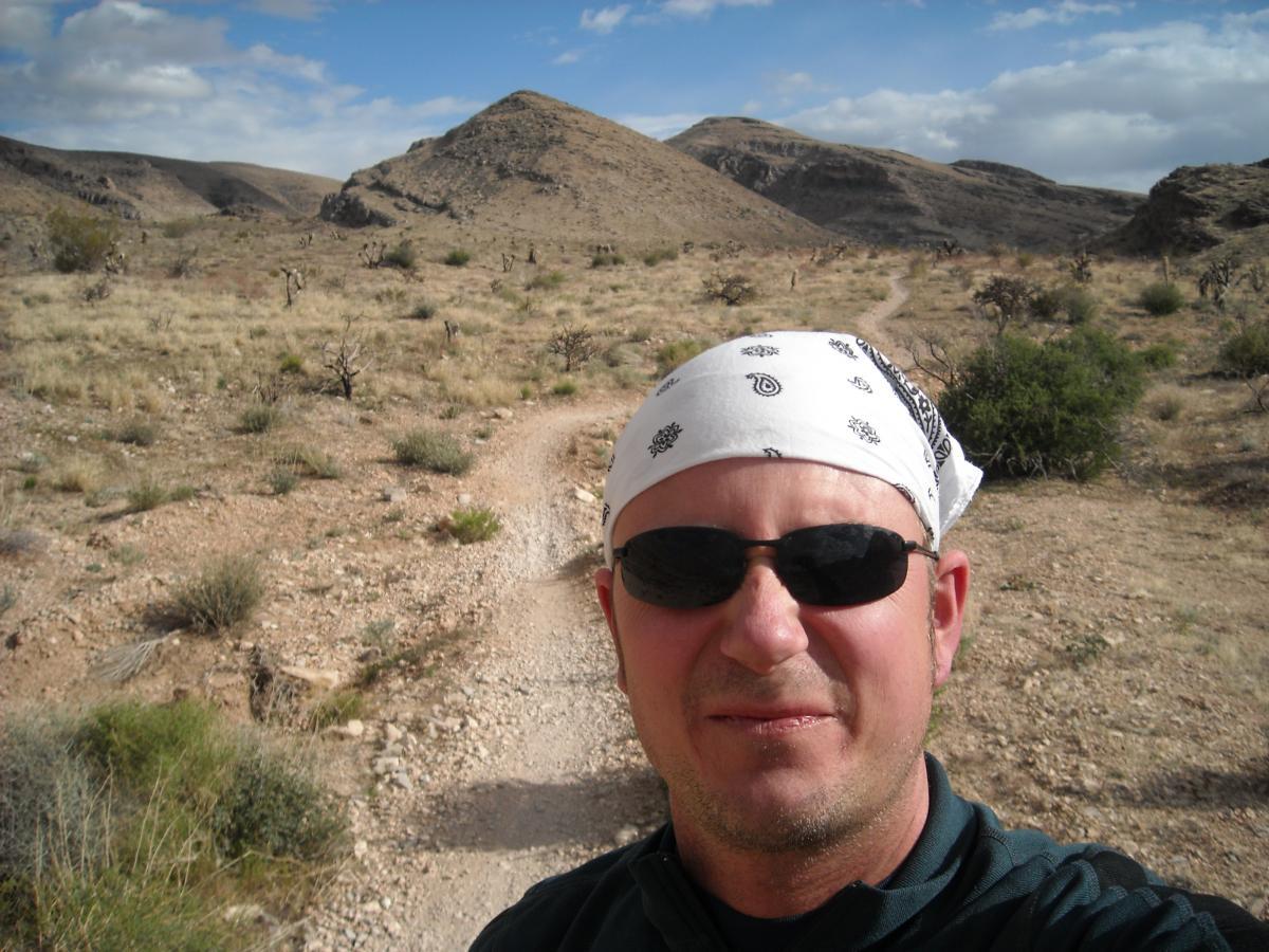 A person in sunglasses and a white bandana stands in a rocky desert landscape, with mountains in the background and a dirt path winding through the dry terrain. The sky is partly cloudy, and sparse vegetation is visible in the foreground. Blue Diamond mountain bike trail.