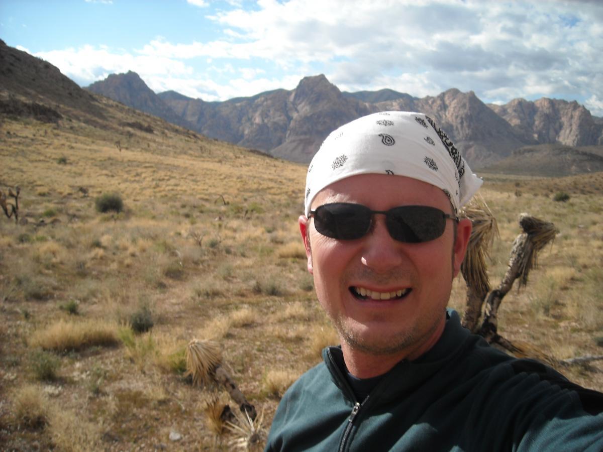Self-portrait of a man wearing sunglasses and a white bandana, smiling in a desert landscape with mountains in the background. The scene features a dry, grassy terrain with sparse vegetation under a partly cloudy sky. Blue Diamond mountain bike trail.