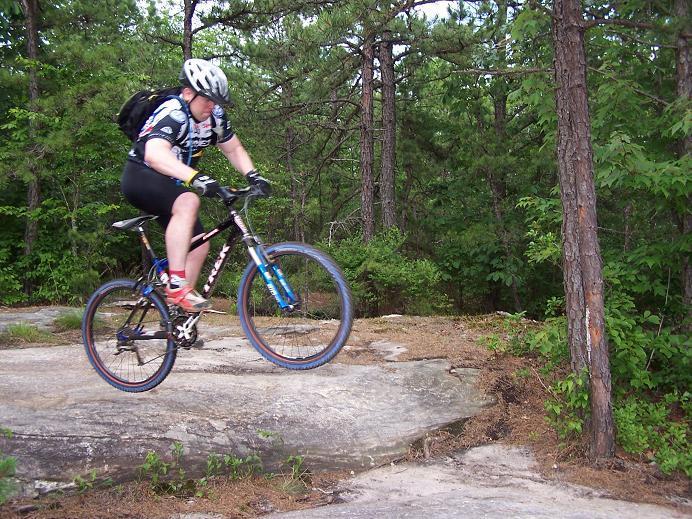 A cyclist in a helmet and biking gear performs a jump on a mountain bike over a rocky terrain, surrounded by trees and greenery. DuPont State Recreational Forest mountain bike trail.