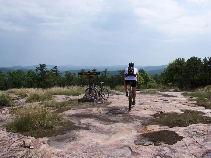 A cyclist riding away on a rocky trail, with a mountain landscape in the background. A second bike is parked next to a sign that reads "BIG ROCK TRAIL." The scene features green trees and a cloudy sky, showcasing an outdoor adventure setting. DuPont State Forest mountain bike trail.