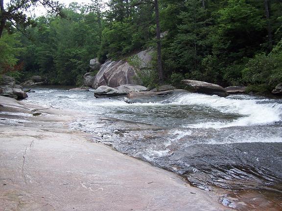 A serene river scene featuring flowing water over smooth rocks, surrounded by dense green foliage and large boulders. The landscape showcases a mix of trees lining the banks, creating a tranquil natural setting. DuPont State Forest mountain bike trail.