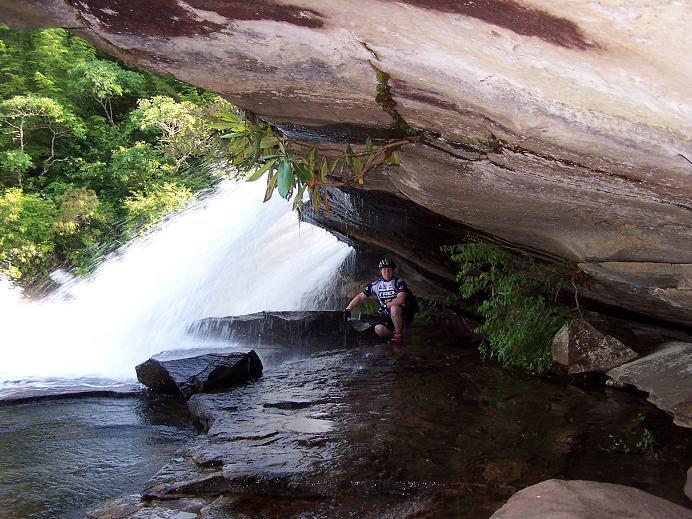 A person wearing a helmet sits on a rocky ledge near a waterfall, surrounded by lush greenery. Water cascades dramatically over the edge, creating a misty atmosphere. The rocky surface and overhanging cliff provide a natural shelter as the person interacts with the water. DuPont State Forest mountain bike trail.