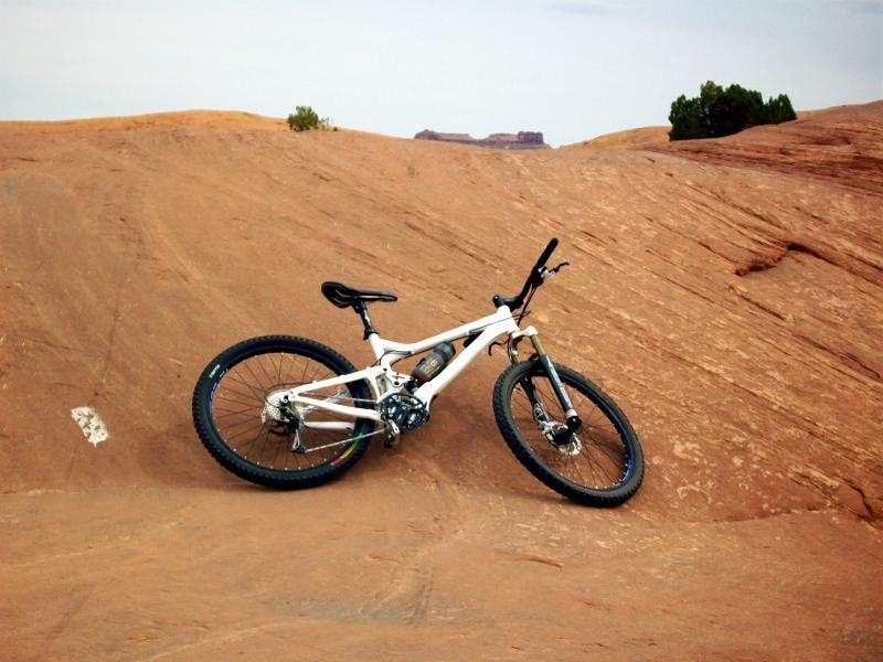 A white mountain bike leaning against a rocky, reddish-brown terrain in an outdoor landscape. The background features subtle hills and sparse vegetation under an overcast sky. Slickrock mountain bike trail.