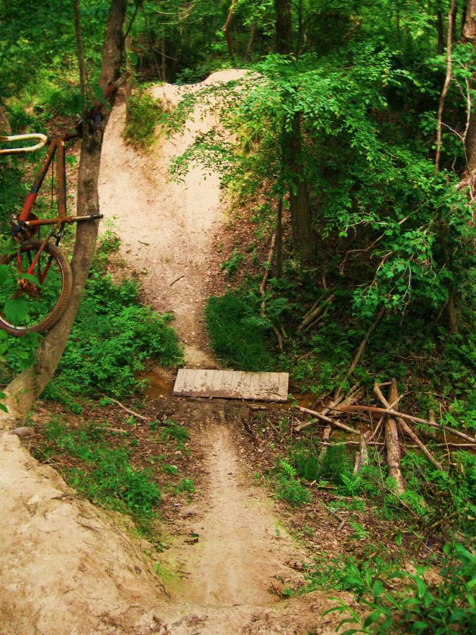 A narrow dirt path winding through a dense green forest, featuring a wooden bridge crossing a small stream. In the foreground, a bicycle frame is hanging from a tree, while the path leads downhill, surrounded by lush vegetation. Comite Trails mountain bike trail.