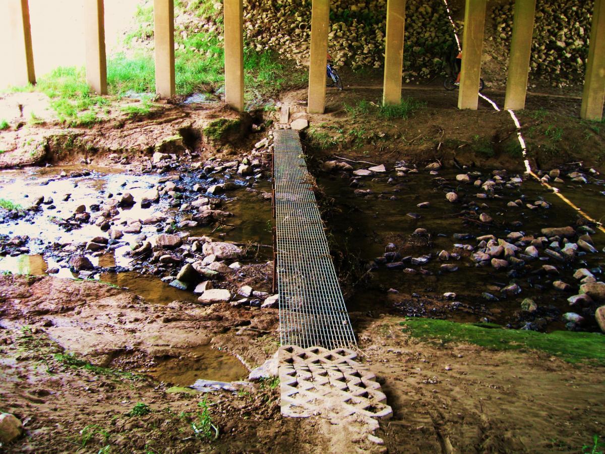 A narrow metal walkway crosses a small stream under a bridge, surrounded by rocky soil and green grass. The bridge's concrete pillars rise in the background, supporting the structure above. Small rocks are scattered along the water's edge, and a few plants are visible growing in the wet soil. Hooper Road mountain bike trail.