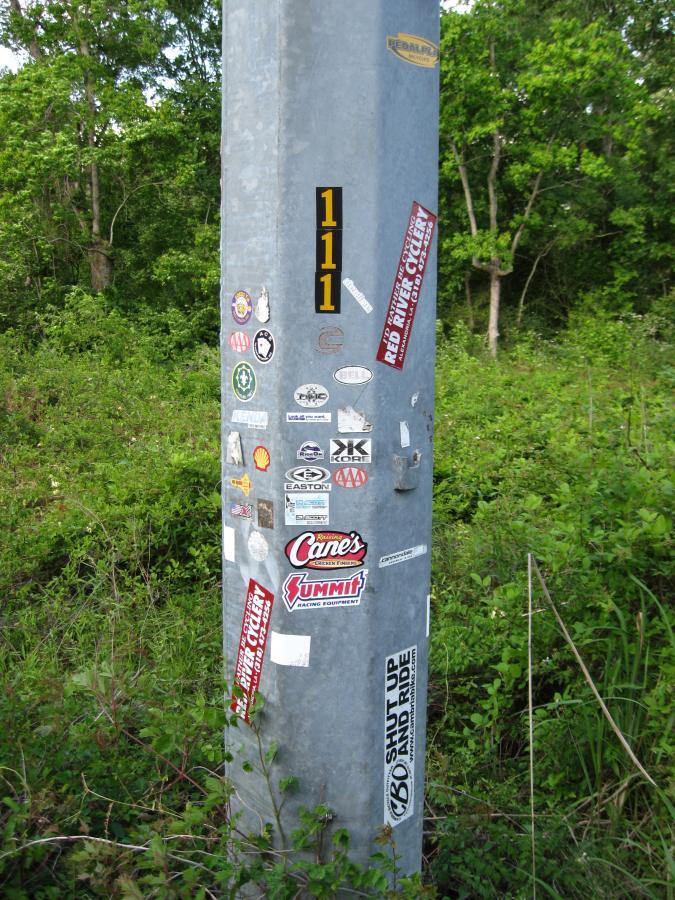 A rusted metal pole covered in various colorful stickers and decals, standing in a grassy area surrounded by trees. The pole displays the number "111" in bold black letters. Stickers include logos from different brands and organizations, along with messages such as "SHUT UP AND RIDE." Hooper Road mountain bike trail.
