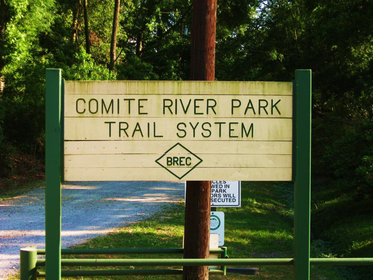 Sign for the Comite River Park Trail System, featuring text indicating park name and management (BREC), surrounded by trees and a gravel path. Comite Trails mountain bike trail.