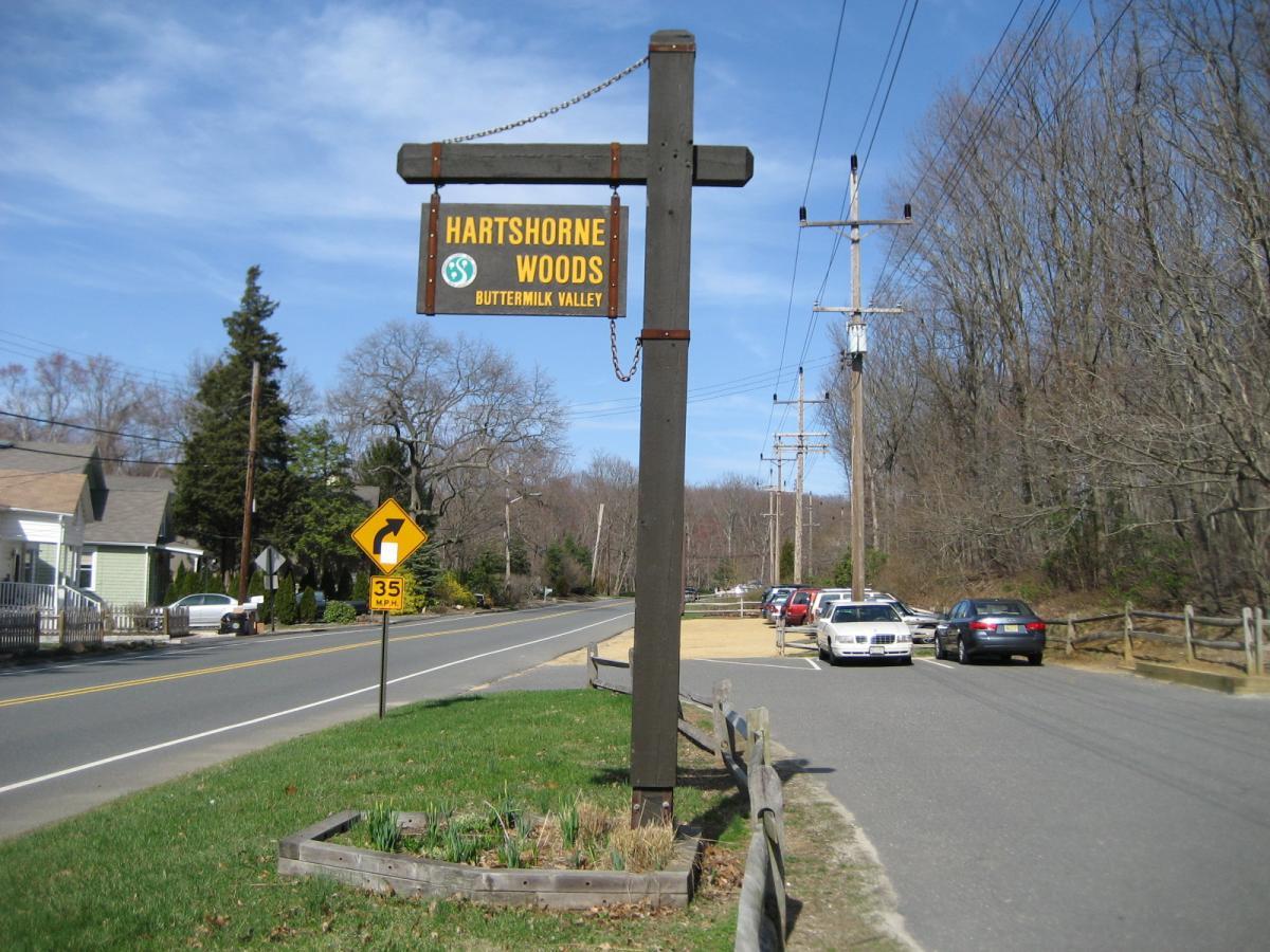 Sign for Hartshorne Woods, Buttermilk Valley, located alongside a road with parked cars, a speed limit sign, and surrounding trees under a clear blue sky. Hartshorne Woods Park mountain bike trail.