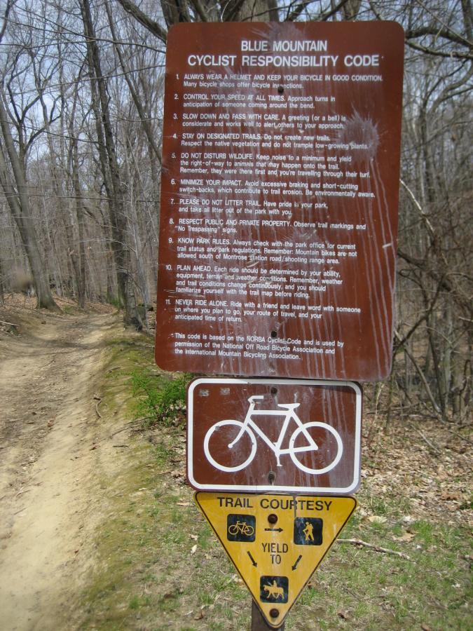 A brown informational sign detailing the "Cyclist Responsibility Code" for Blue Mountain, which includes guidelines for safe biking practices, trail etiquette, and respect for wildlife. Below it, another sign indicates "Trail Courtesy" with symbols for yielding to other trail users. The background features a dirt path through a wooded area. Blue Mountain Reservation mountain bike trail.