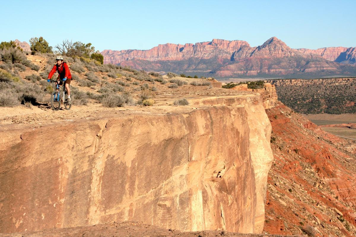 A person riding a mountain bike along a rocky cliff edge with a vast landscape and mountain range in the background. The scene features dry terrain, sparse vegetation, and clear blue skies. Gooseberry Mesa mountain bike trail.