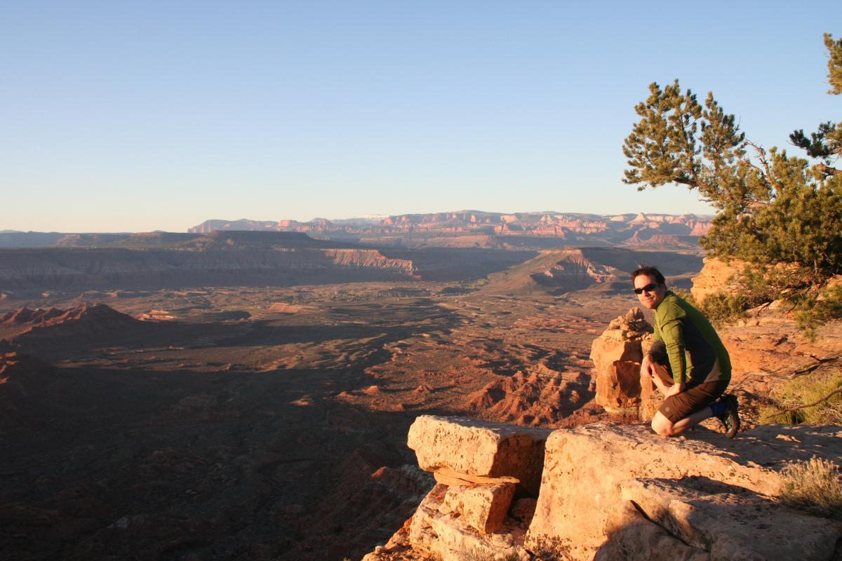 A person kneels on a rocky outcrop, overlooking a vast desert landscape with layered mountains and valleys illuminated by the golden light of sunrise. The sky is clear and blue, contrasting with the warm tones of the rugged terrain. A tree is visible in the foreground, adding to the natural scenery. Gooseberry Mesa mountain bike trail.
