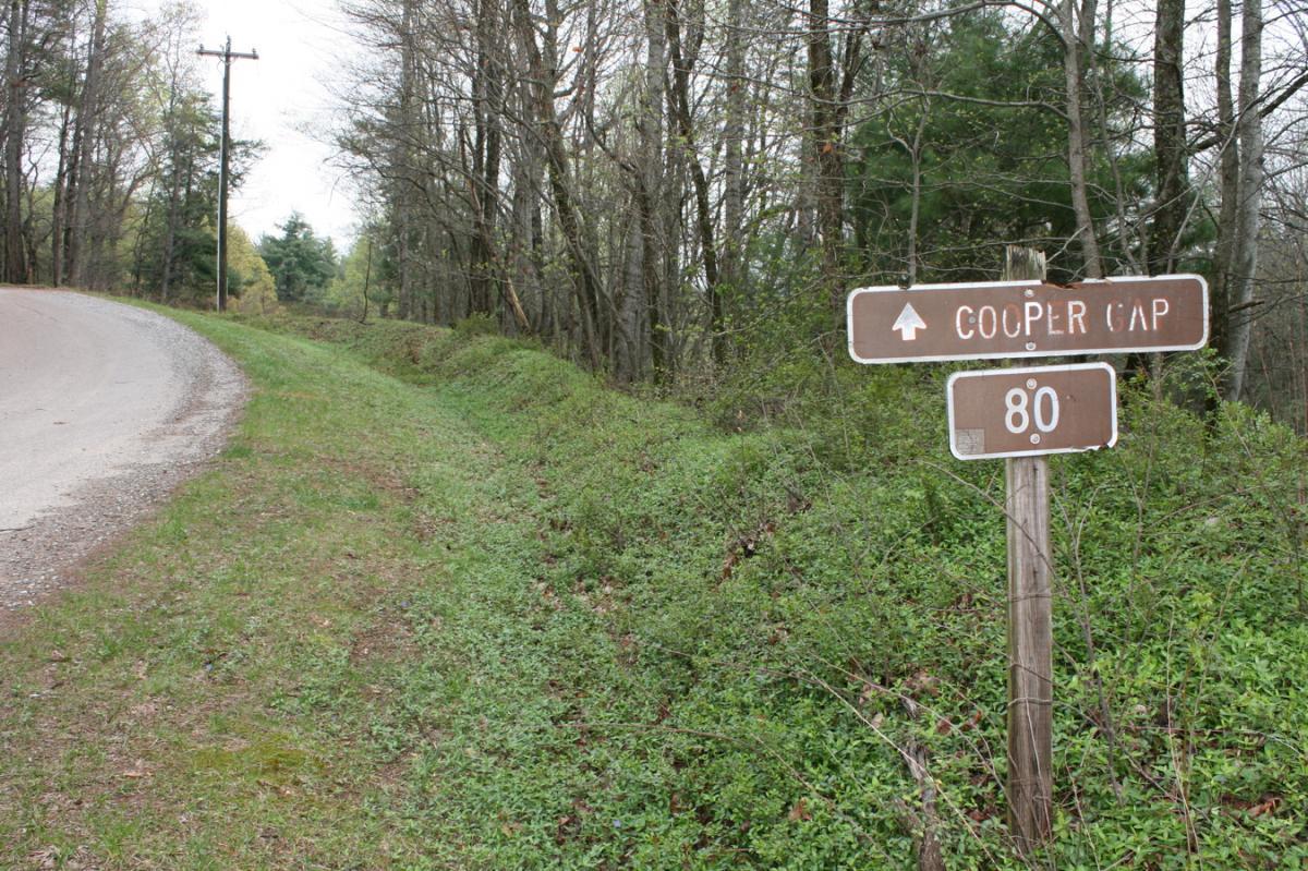 A roadside sign indicating "Cooper Gap" with an arrow pointing up and a distance marker showing "80". The background features a winding gravel road lined with trees and greenery, suggesting a rural or wooded area. Winding Stairs Loop mountain bike trail.