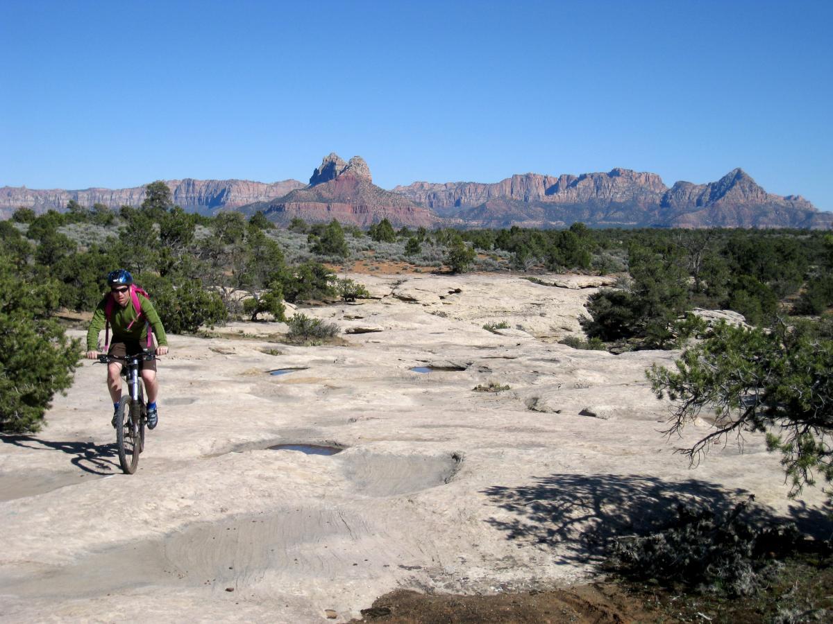A mountain biker rides along a rocky trail with a clear blue sky above. The landscape features sparse vegetation and a backdrop of rugged mountains in the distance, showcasing natural terrain typical of an outdoor adventure setting. Gooseberry Mesa mountain bike trail.