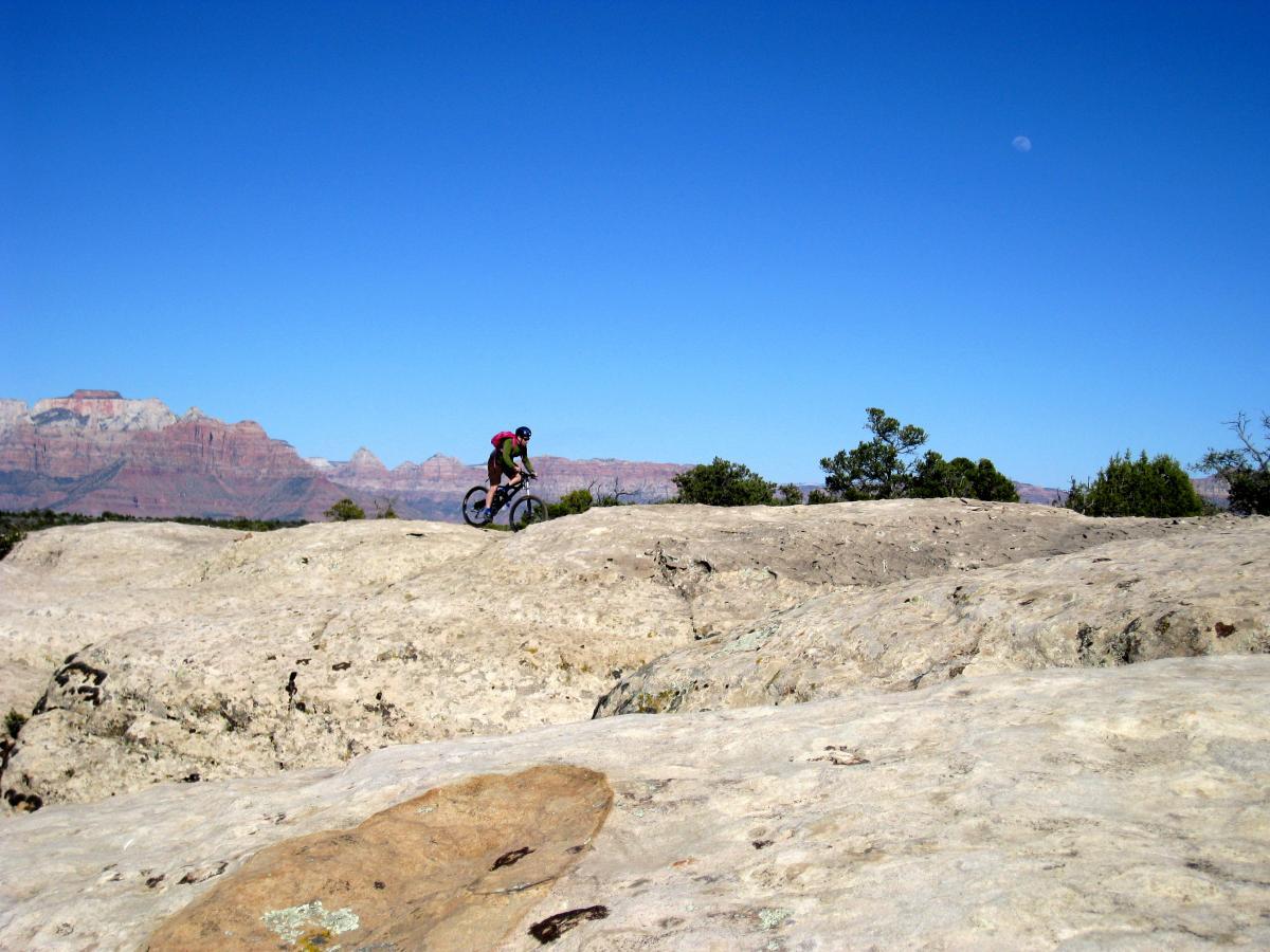 A mountain biker navigating over rocky terrain with a backdrop of red rock formations and a clear blue sky. The scene captures the essence of outdoor adventure in a rugged landscape. Gooseberry Mesa mountain bike trail.