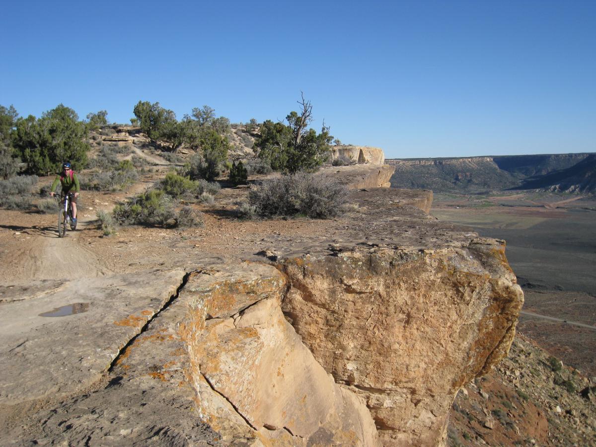 A cyclist riding on a gravel trail near a cliff edge, surrounded by sparse vegetation and rocky terrain under a clear blue sky. The landscape features a vast view of distant hills and valleys. Gooseberry Mesa mountain bike trail.