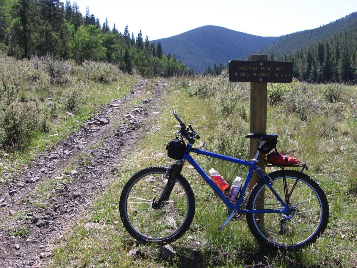 A blue mountain bike rests beside a wooden trail sign indicating "Razor CR TR NO 487" and "COLO TR/CONST TR 65." The scene features a rugged dirt path leading into a forested area, with green foliage and distant mountains in the background under a clear blue sky. Dutchman Creek / #490 mountain bike trail.
