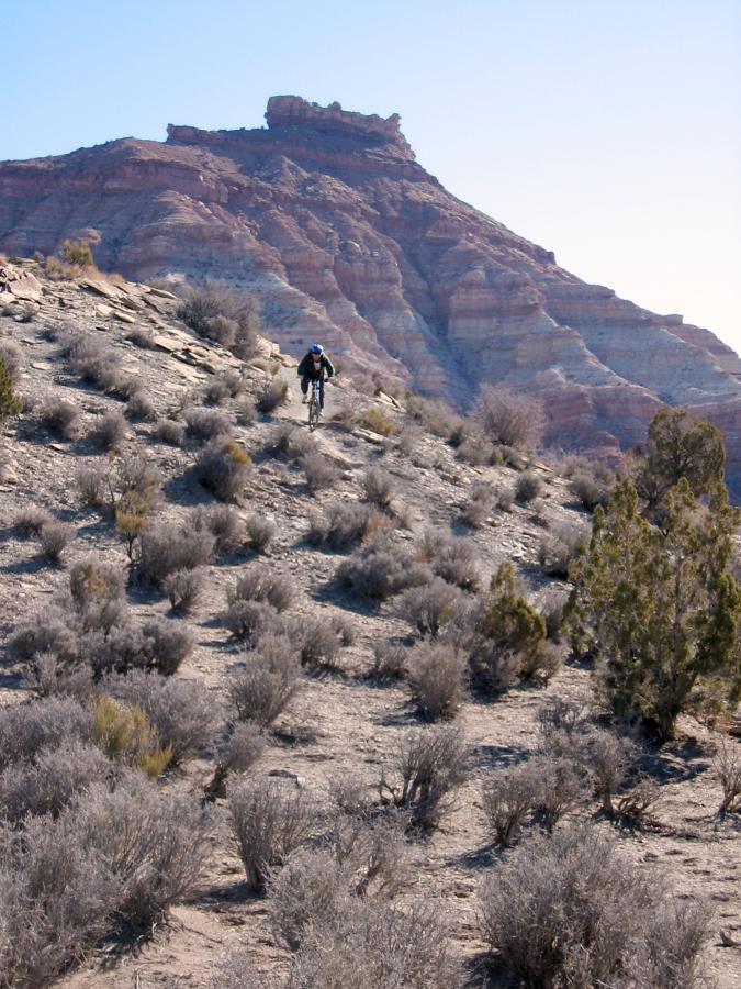 A cyclist rides along a rocky terrain with sparse vegetation, set against a backdrop of a prominent layered mountain under a clear blue sky. J.E.M. Trail mountain bike trail.