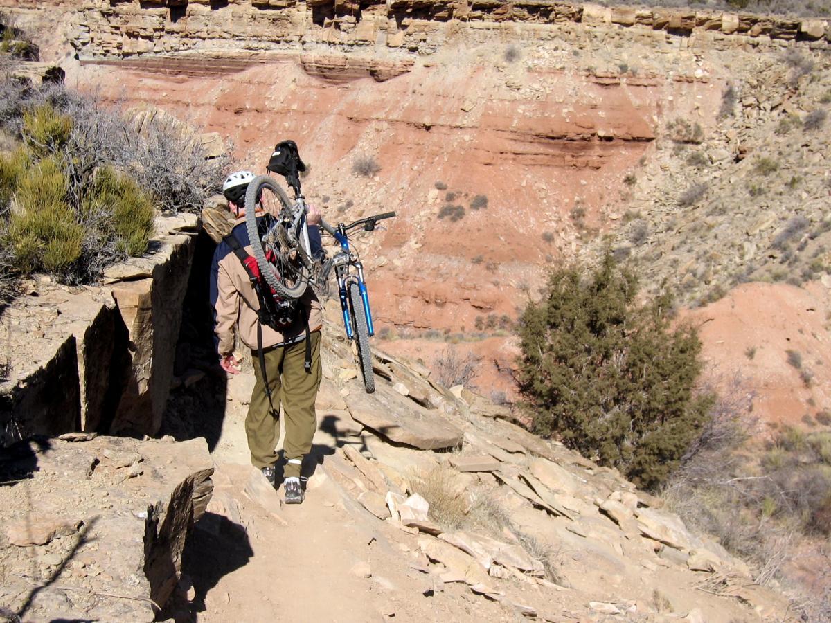A person wearing a bicycle helmet and carrying a mountain bike over their shoulder is walking along a rocky trail in a rugged landscape. The background features layered red rock formations and sparse vegetation typical of a desert environment. J.E.M. Trail mountain bike trail.