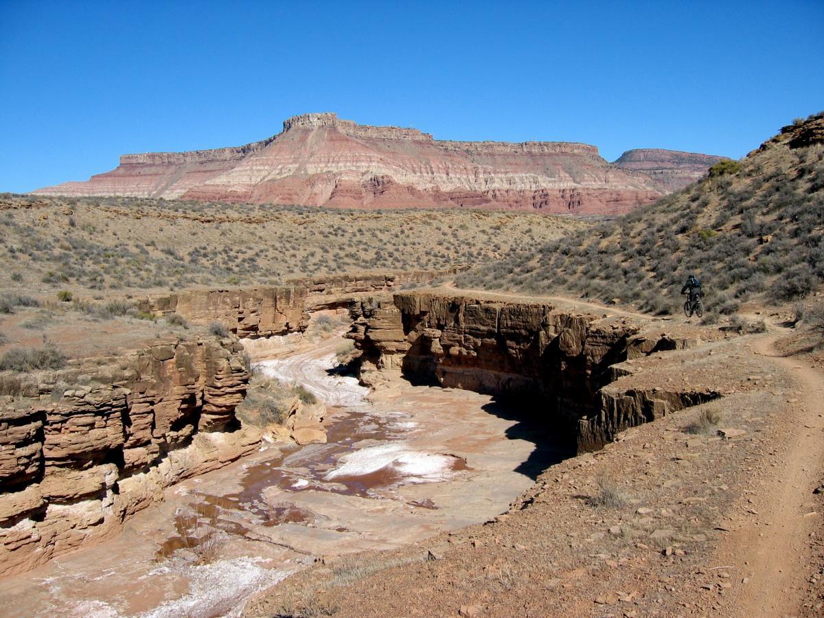 A scenic landscape featuring a rugged canyon with a river running through it, surrounded by arid terrain and sparse vegetation. In the background, a prominent red rock formation rises under a clear blue sky. A solitary mountain biker is seen on a dirt trail along the canyon's edge. J.E.M. Trail mountain bike trail.