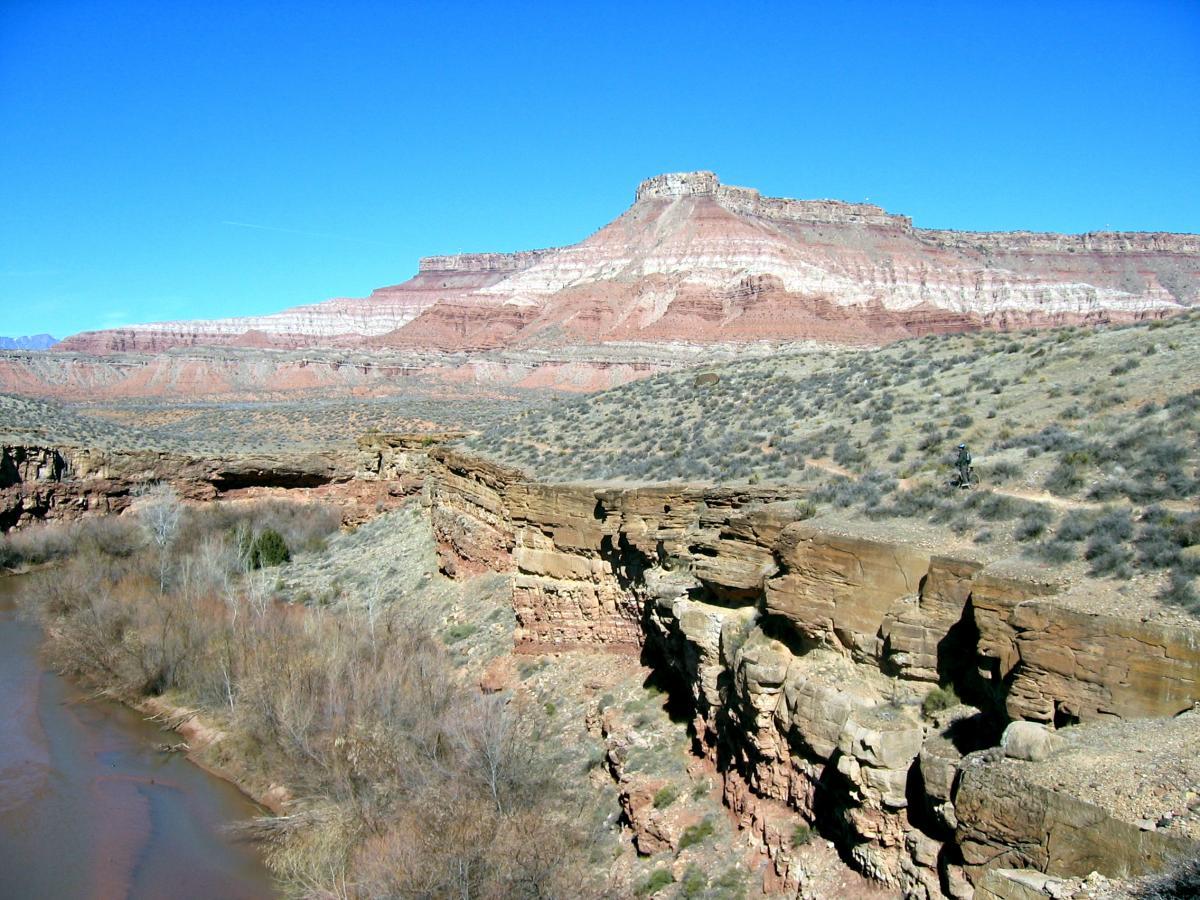 A scenic landscape featuring a rugged canyon with layered rock formations under a clear blue sky. A river winds through the canyon, surrounded by sparse vegetation and rocky cliffs, showcasing the natural beauty of the terrain. J.E.M. Trail mountain bike trail.
